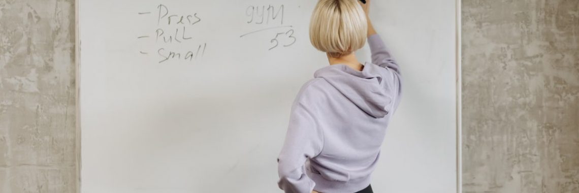 Female writing workout schedule on whiteboard in gym setting.